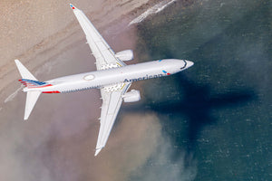 An American Airlines passenger jet is flying over a body of water, with its shadow visible on the water below.