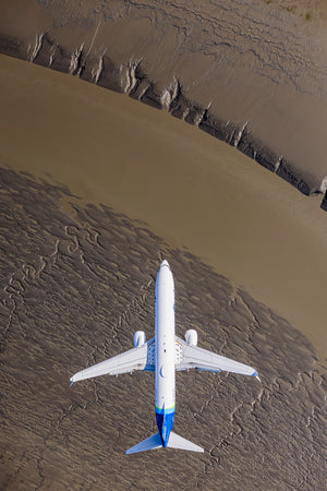 An aerial view of a white airplane with blue accents flying over a dry, cracked landscape.