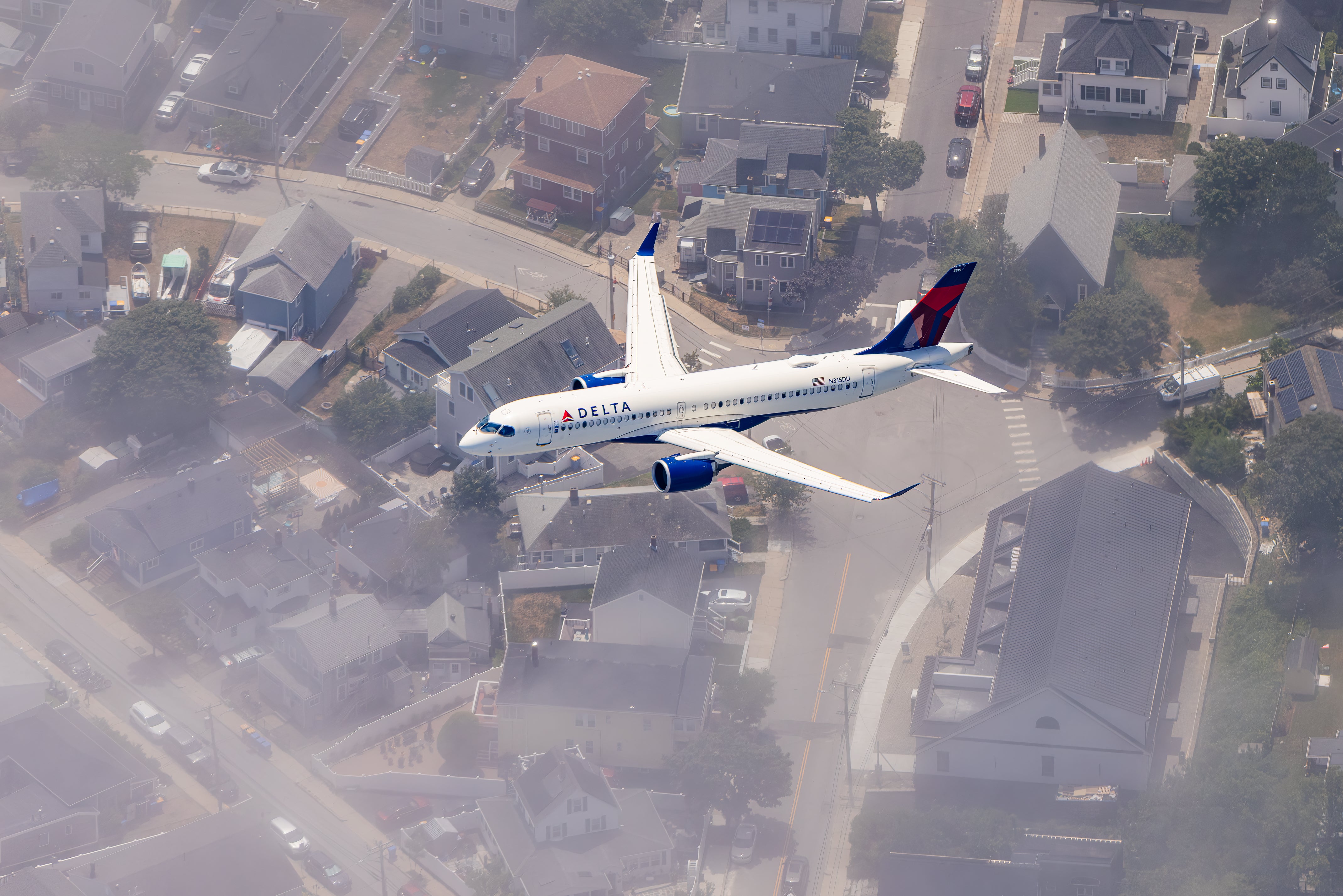 An aerial view of a large commercial airplane flying over a residential neighborhood with houses and cars visible below.