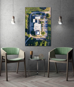 a modern living room with a large wall-mounted painting depicting an aerial view of a building surrounded by greenery. The room is furnished with two green chairs and a small table, creating a cozy and inviting atmosphere.