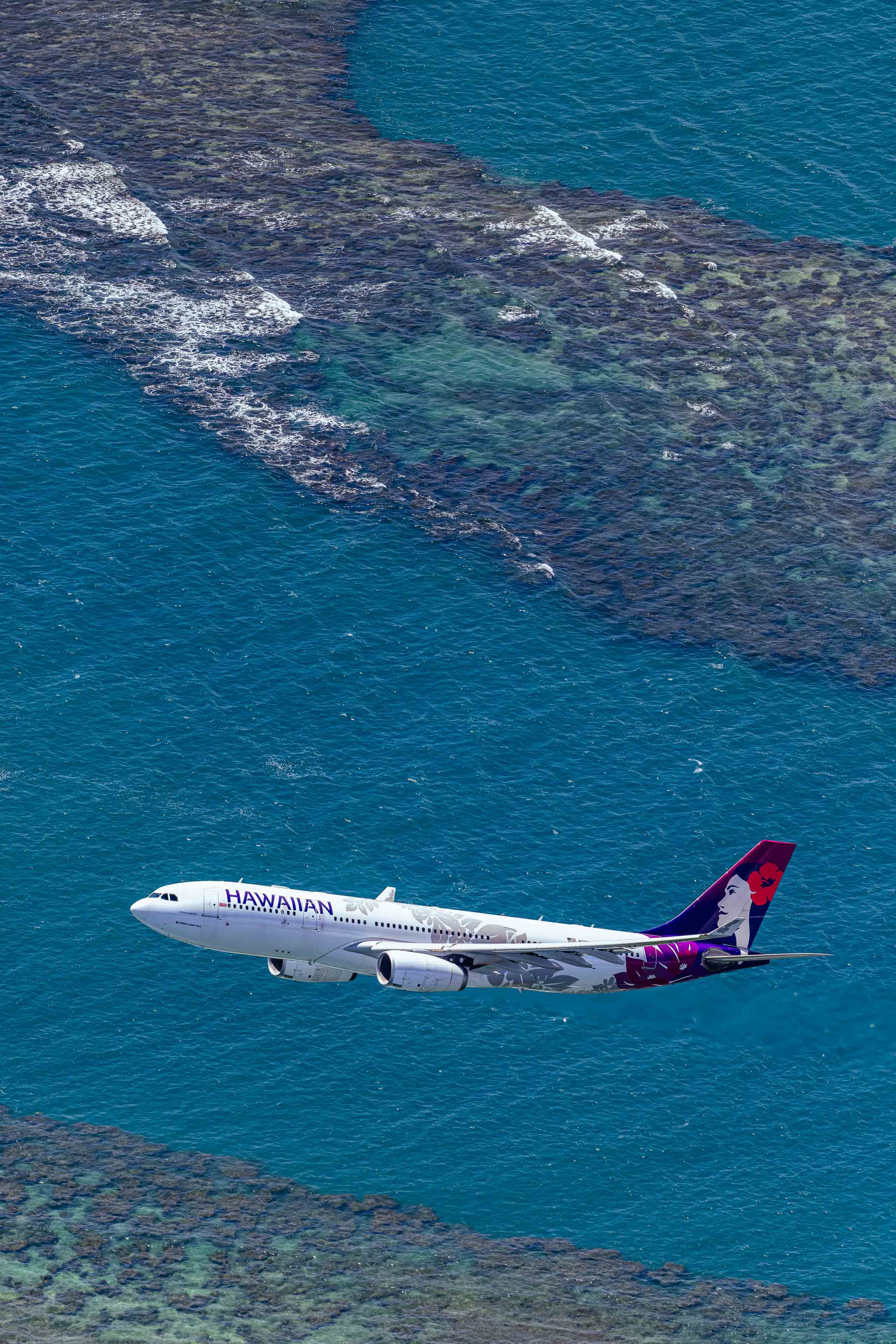 An aerial view of a Hawaiian Airlines passenger jet flying over a coral reef and the ocean.