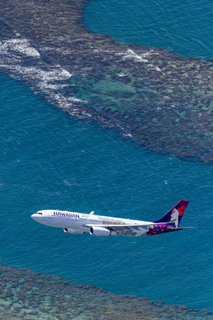 An aerial view of a Hawaiian Airlines passenger jet flying over a coral reef and the ocean.