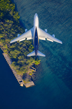 An aerial view of a large commercial airplane flying over a body of water, with a wooden dock and lush vegetation on the shoreline below.