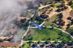 An aerial view of a blue and white airplane flying over a grassy area with trees and buildings, surrounded by a misty landscape.