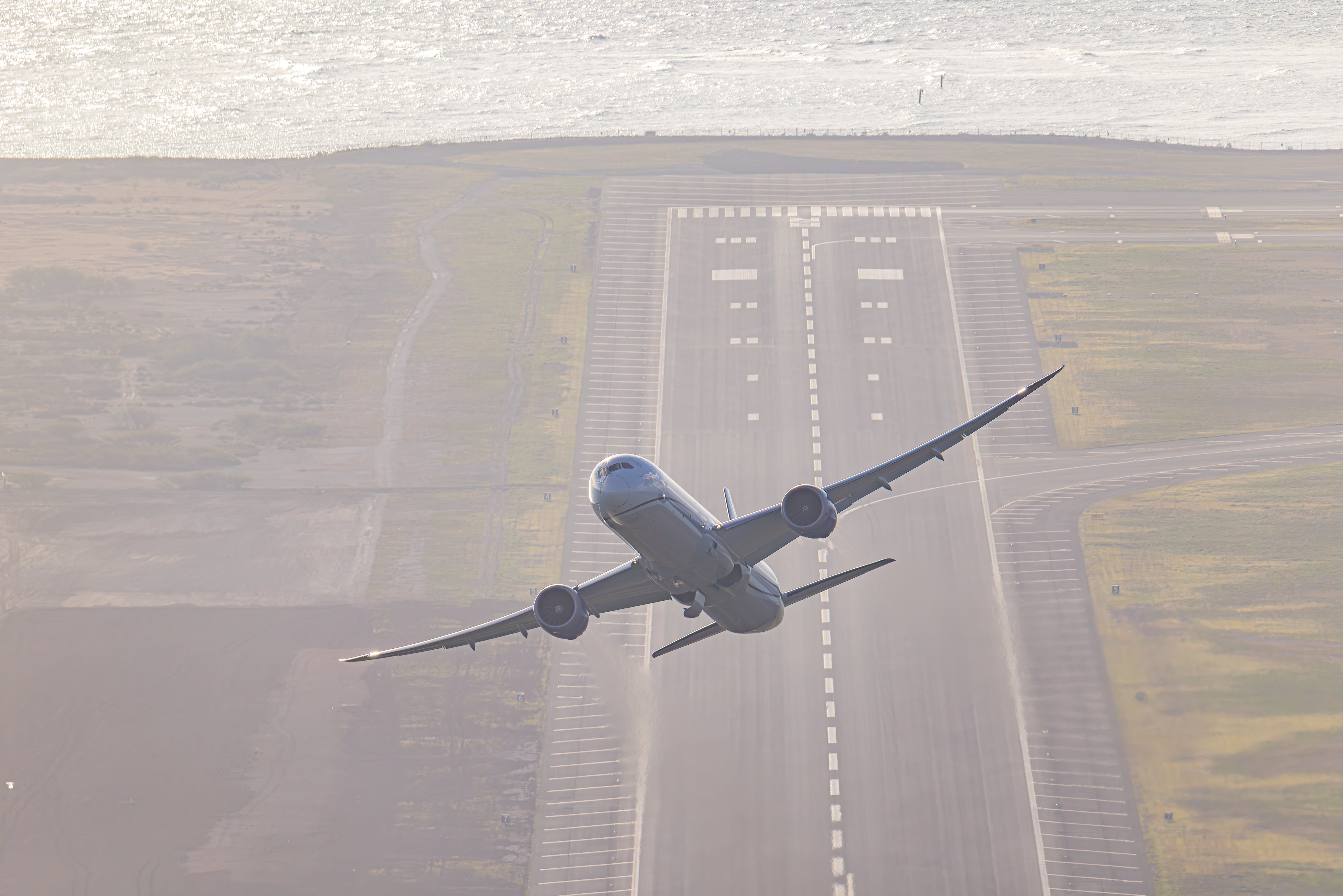 An airplane is flying low over a runway, with the ocean visible in the background.
