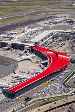 An aerial view of an airport with multiple airplanes parked at the terminal and a large red curved structure, likely a terminal building, prominently featured.
