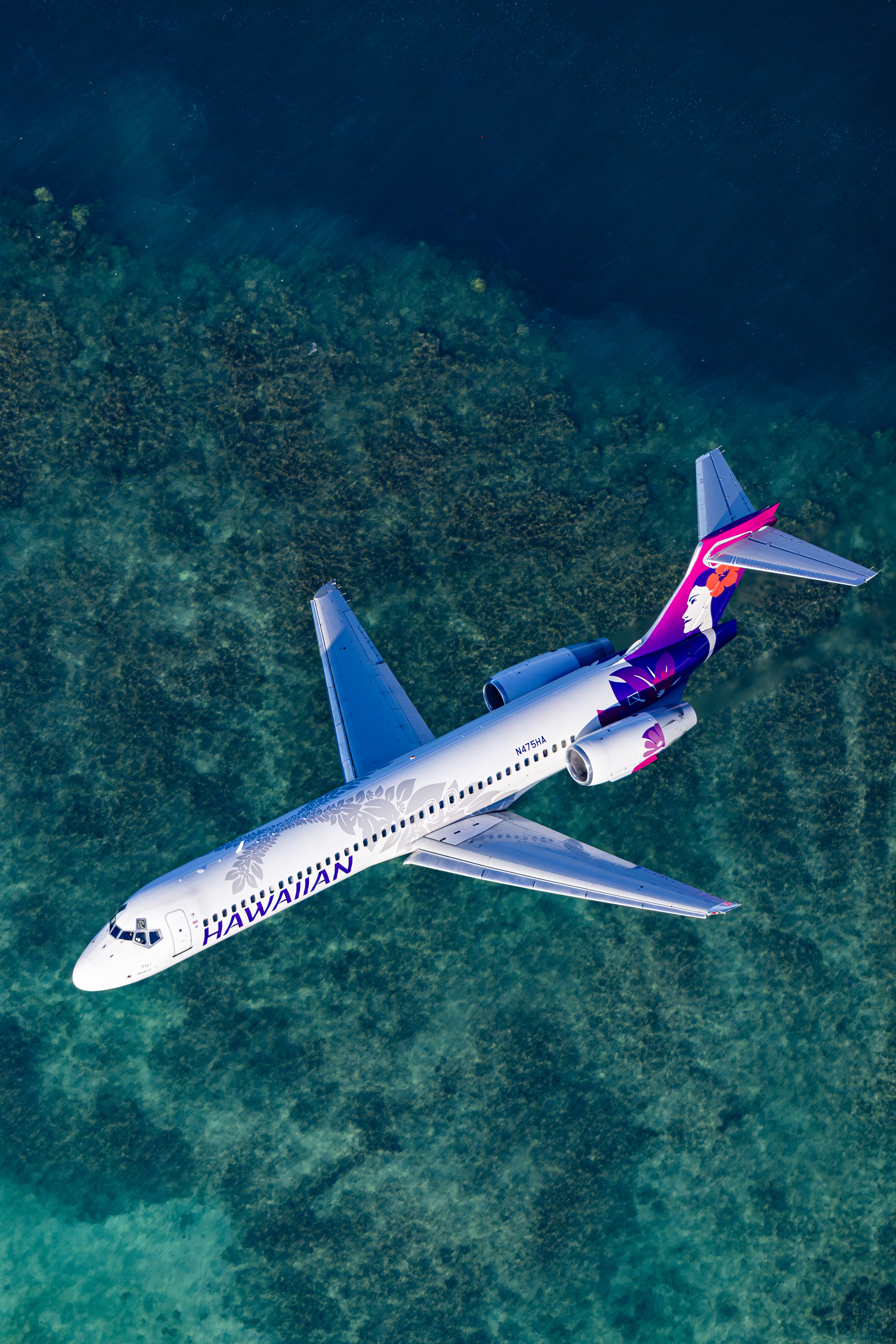 An aerial view of a Hawaiian Airlines passenger jet flying over a body of water, with the ocean visible below the plane.