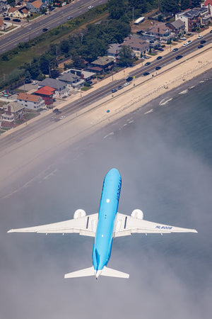 An aerial view of a blue and white airplane flying over a coastal area with houses and a beach visible below.