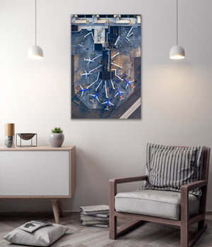 a modern living room with a large wall-mounted painting depicting an airport terminal with multiple airplanes. The room has a minimalist design with a wooden chair, a white cabinet, and a stack of books on the floor.