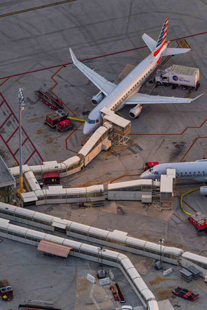 An aerial view of an airport tarmac with a large commercial airplane parked at a gate, surrounded by various ground support vehicles and equipment.