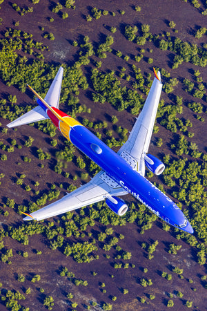 An aerial view of a Southwest Airlines passenger jet flying over a landscape with green vegetation and brown terrain.