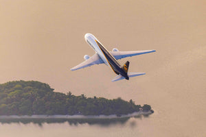 An aerial view of a large commercial airplane flying over a body of water, with a lush green island visible in the distance.