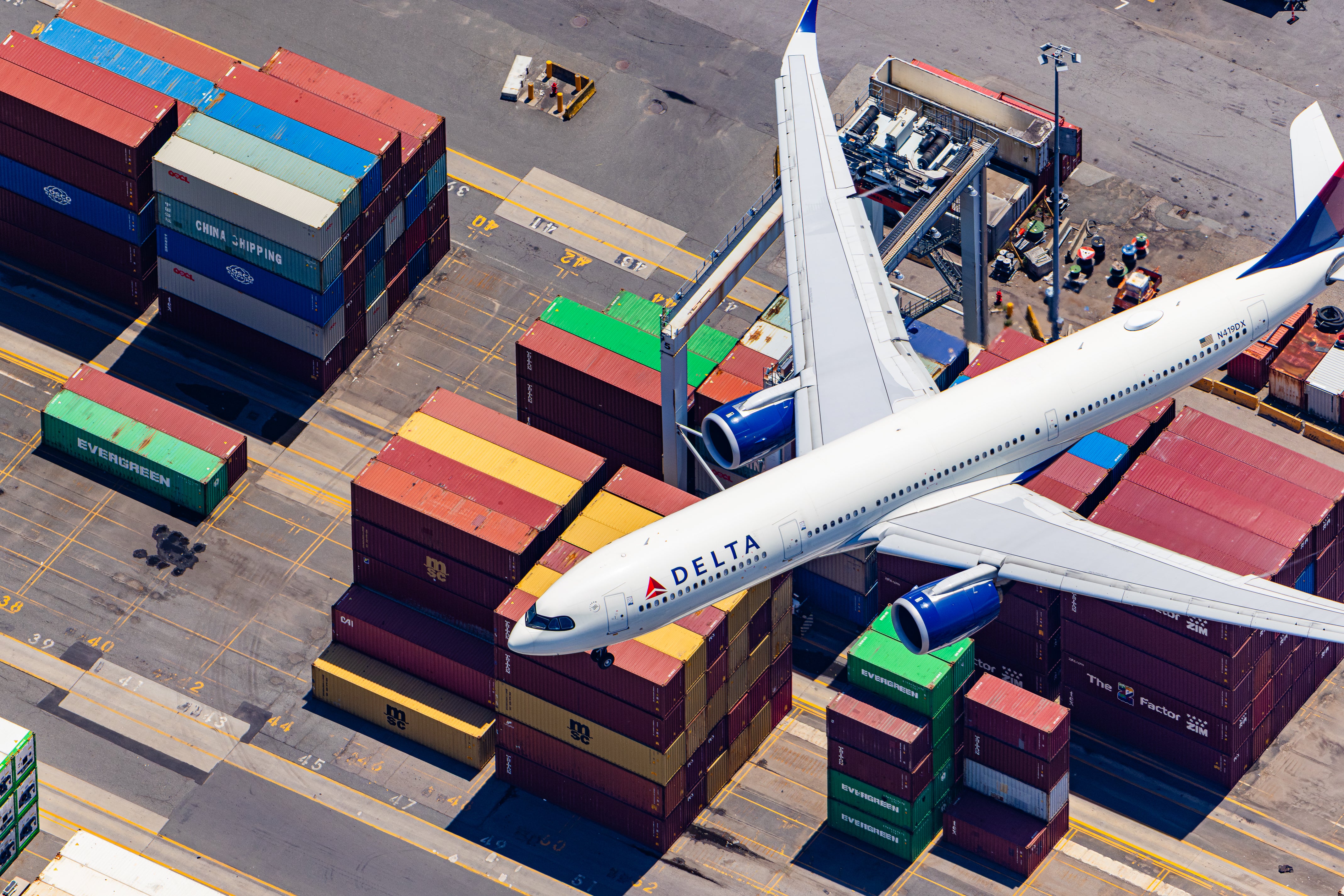 An aerial view of a Delta Airlines plane flying over a busy cargo area with numerous shipping containers stacked around it.