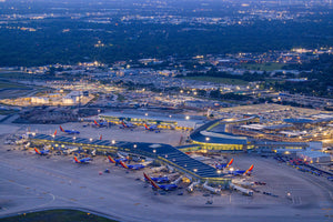An aerial view of a bustling airport at night, with numerous airplanes parked at the terminal and various airport buildings and infrastructure visible in the background.