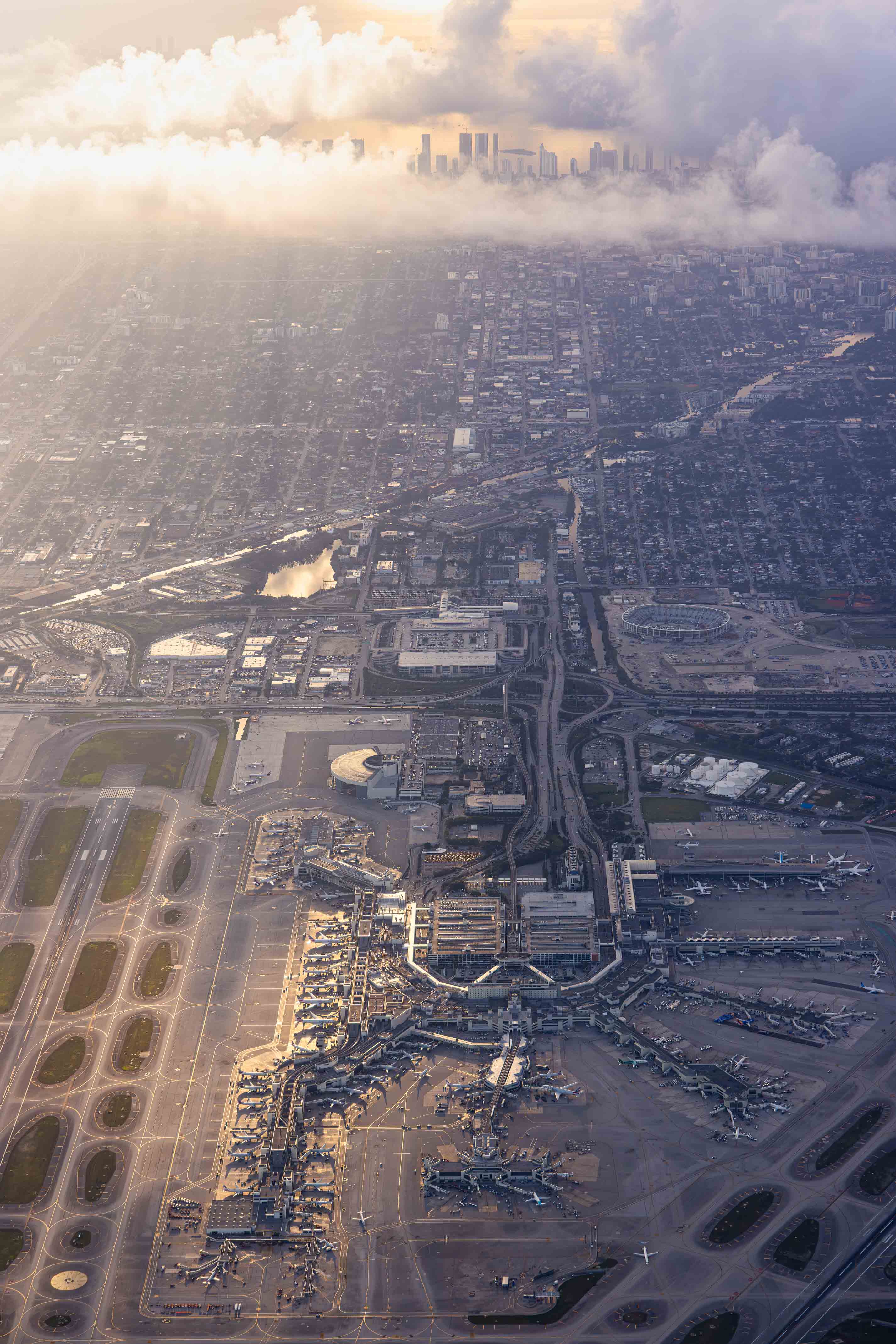 An aerial view of a bustling city with a large airport in the center, surrounded by numerous buildings and roads. The sky above is filled with clouds, creating a dramatic backdrop for the cityscape.
