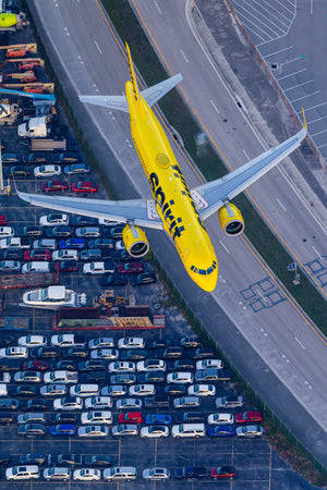 An aerial view of a yellow airplane flying over a parking lot filled with cars.