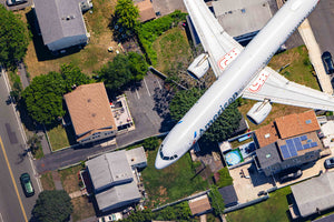 An aerial view of a white airplane with red and blue markings flying over a residential neighborhood with houses and trees.