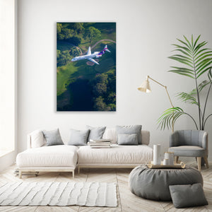 a large white sofa with a coffee table in a modern living room, with a large window allowing natural light to fill the space. A framed photograph of a purple and white airplane flying over a lush green landscape is displayed on the wall above the sofa.
