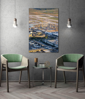 a modern living room with a large wall-mounted painting depicting an aerial view of an airport and its surrounding area. The painting is hung above a small table with two green chairs placed in front of it. The room has a minimalist and contemporary design, with a gray concrete wall and a wooden floor.