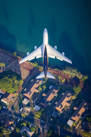 An aerial view of a large passenger jet flying over a residential area with houses and palm trees.