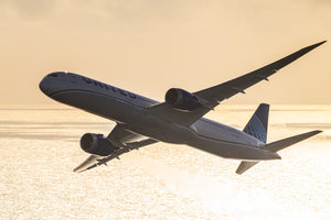 A large commercial airplane is flying over a body of water, with the sun setting in the background.