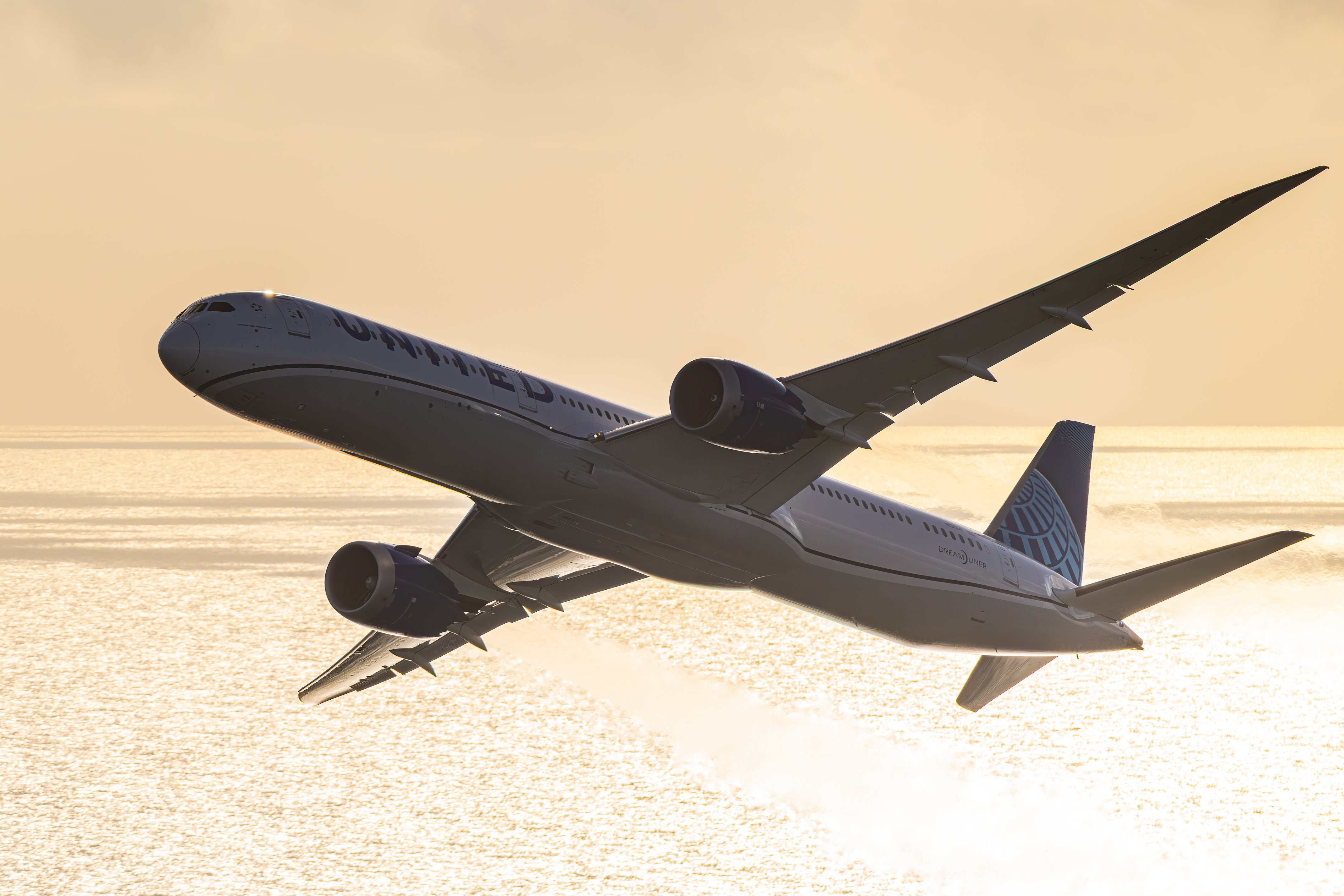 A large commercial airplane is flying over a body of water, with the sun setting in the background.