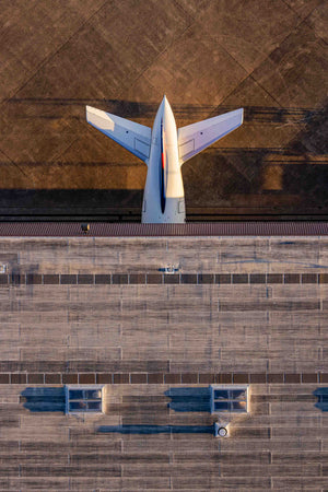 An aerial view of a large white airplane parked on a concrete surface, with a building and a runway visible in the background.