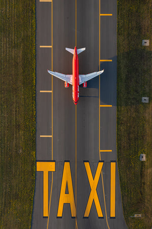 An aerial view of a red airplane taxiing on a runway with the word "TAXI" painted on the ground.