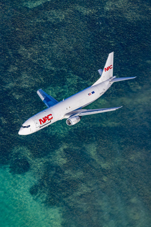An aerial view of a large commercial passenger jet flying over a body of water, with the jet's tail and nose prominently visible.