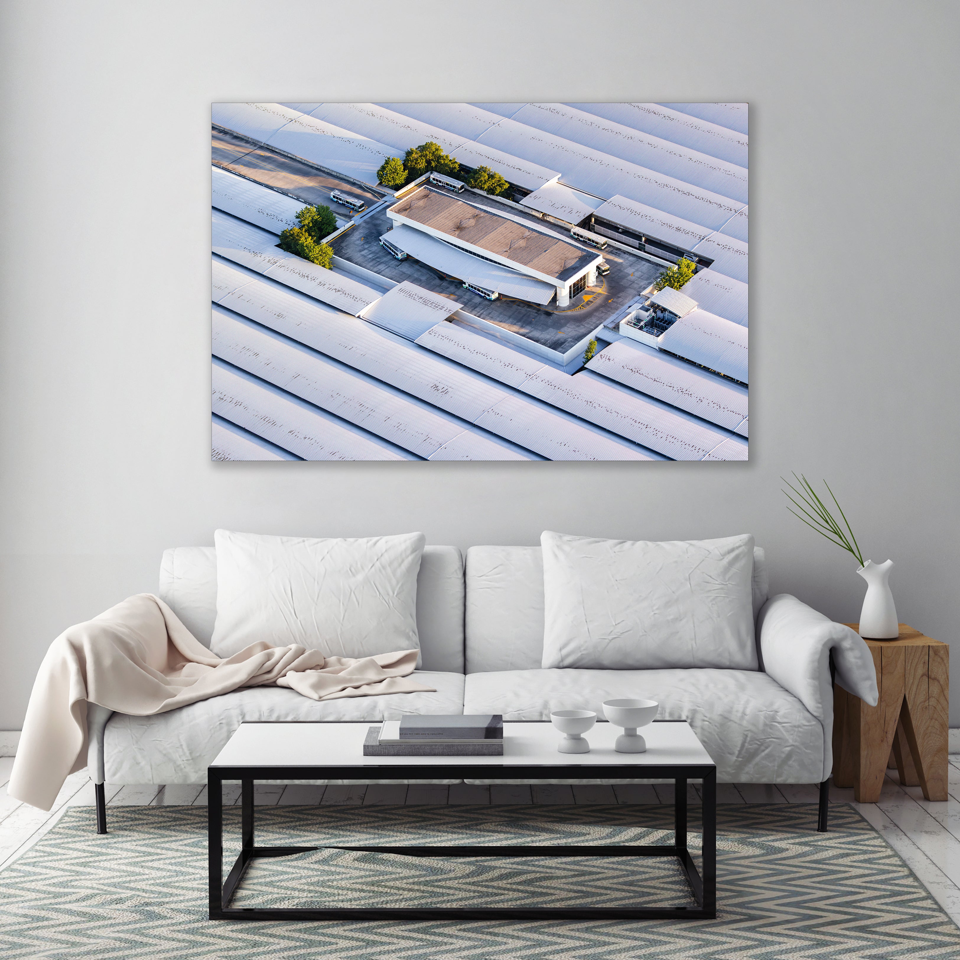 a large white sofa with two white pillows, a black coffee table, and a wooden side table with a white vase. Above the sofa, there is a large framed picture of a building with a flat roof and a small garden area.