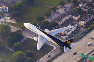 An aerial view of a large white and black cargo plane with the word "Worldwide Services" on its side, flying over a city with buildings, roads, and trees below.