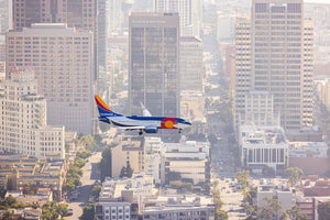 A commercial airplane with a red and white body and blue tail is flying over a cityscape with tall buildings and a hazy sky.