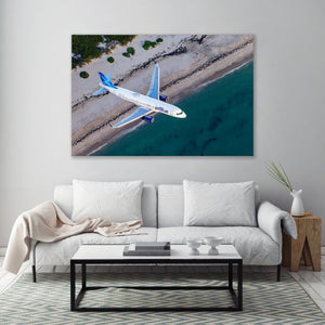 a large, white airplane flying over a beach with a clear blue sky and ocean in the background.