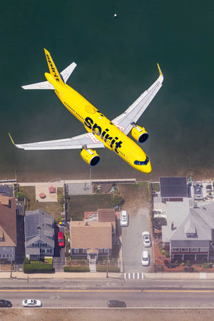 An aerial view of a yellow Spirit Airlines jet flying over a residential area with houses and cars below.