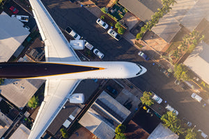 An aerial view of a large commercial airplane flying over a city with buildings, cars, and trees below.