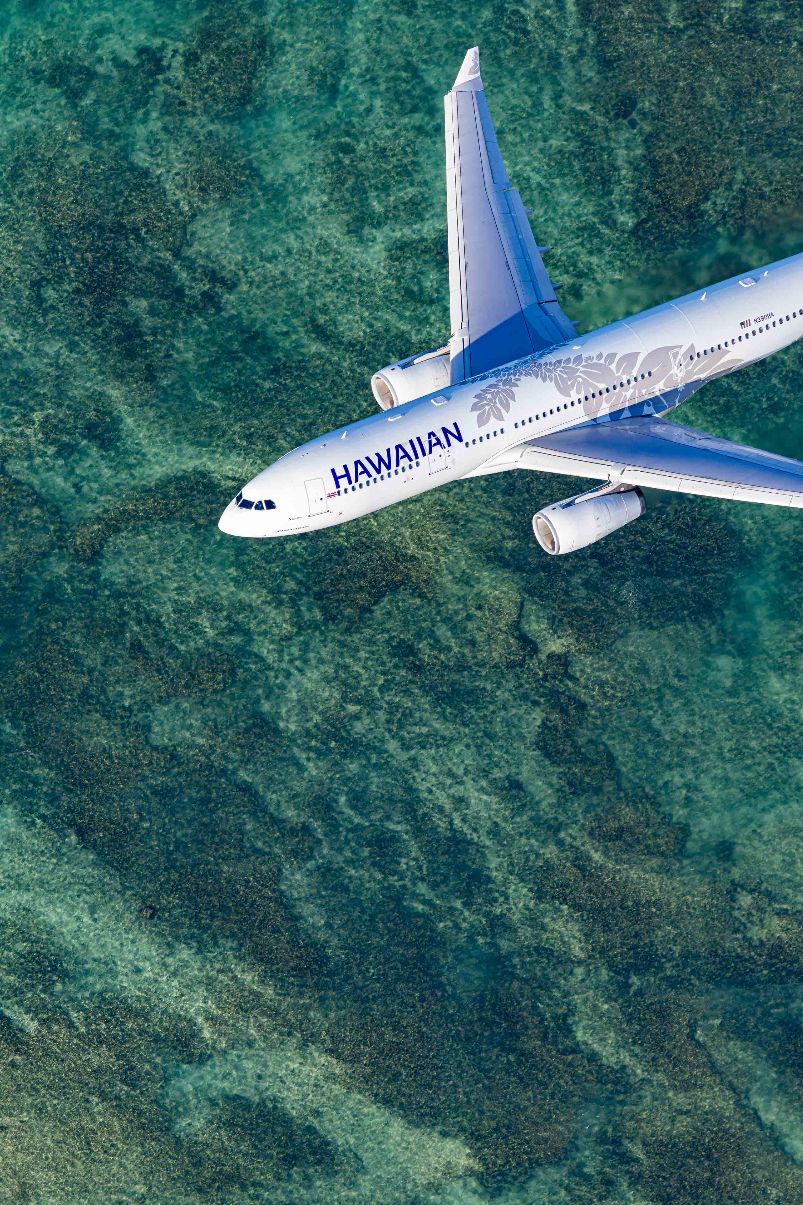 An aerial view of a Hawaiian Airlines passenger jet flying over a body of water with a greenish-blue color.