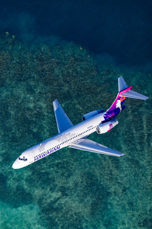 An aerial view of a Hawaiian Airlines passenger jet flying over a body of water, with the ocean visible below the plane.