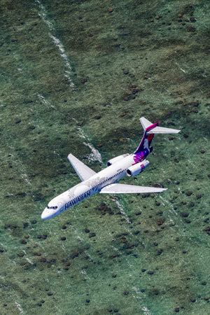An aerial view of a white airplane with purple and pink accents flying over a grassy field.