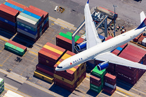 An aerial view of a Delta Airlines plane flying over a busy cargo area with numerous shipping containers stacked around it.