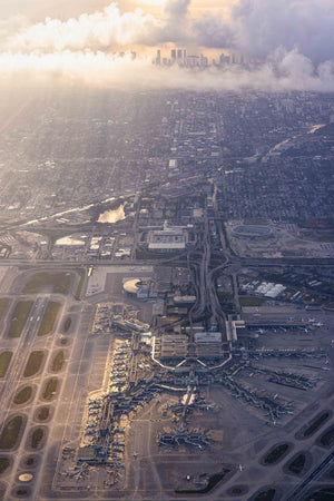 An aerial view of a bustling city with a large airport in the center, surrounded by numerous buildings and roads. The sky above is filled with clouds, creating a dramatic backdrop for the cityscape.