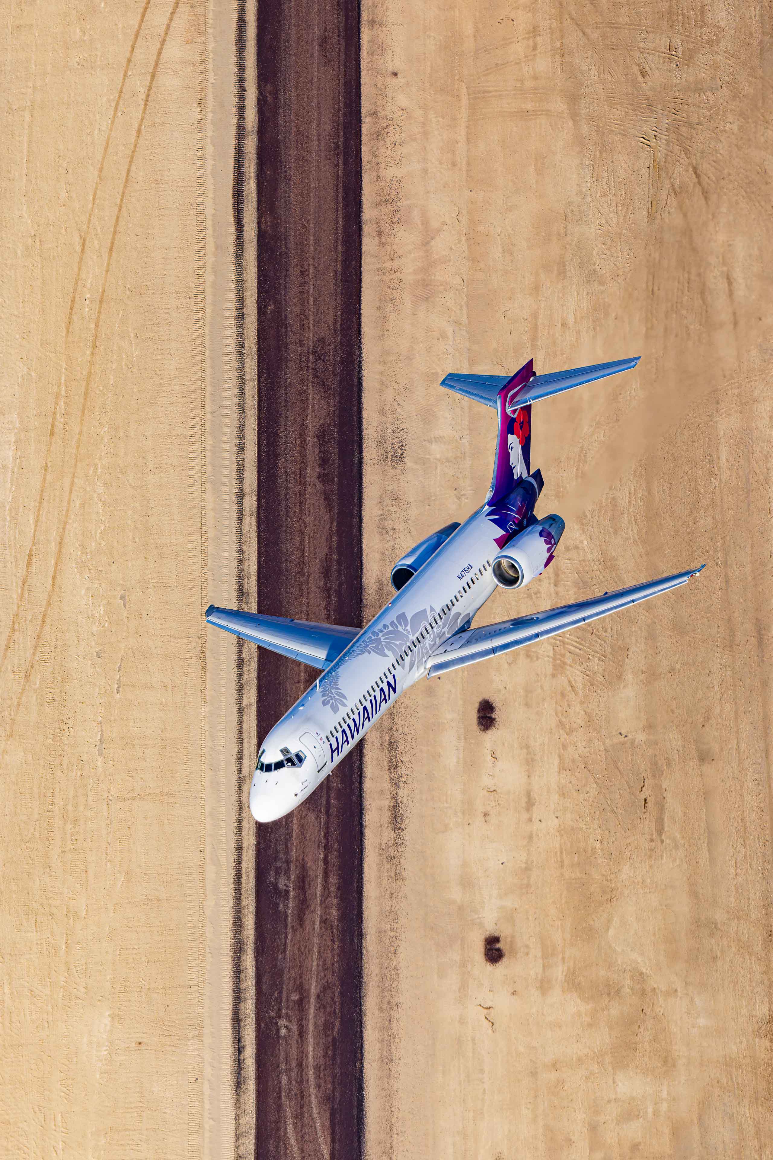 An aerial view of a Hawaiian Airlines passenger jet flying over a runway.